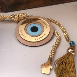 May include: A gold-colored wooden circle with a blue evil eye charm and the words "TUXH", "YGEIA", "EUTUXIA", and "AGAPI" inscribed around the eye. The circle is attached to a gold rope with a tassel and a small gold house charm.