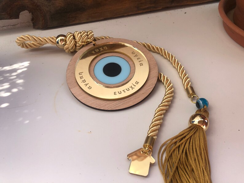 May include: A gold-colored wooden circle with a blue evil eye charm and the words "TUXH", "YGEIA", "EUTUXIA", and "AGAPI" inscribed around the eye. The circle is attached to a gold rope with a tassel and a small gold house charm.
