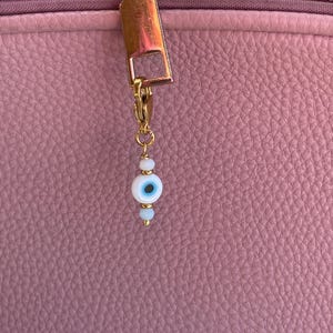 Pu&ograve; includere: Un tirazip dorato con un piccolo ciondolo con un design a occhio blu e bianco. Il ciondolo &egrave; fatto di piccole perline e pende da una chiusura dorata. Lo sfondo &egrave; una superficie rosa strutturata.