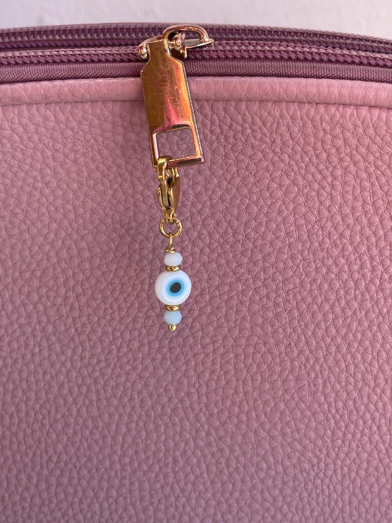 Pu&ograve; includere: Un tirazip dorato con un piccolo ciondolo con un design a occhio blu e bianco. Il ciondolo &egrave; fatto di piccole perline e pende da una chiusura dorata. Lo sfondo &egrave; una superficie rosa strutturata.