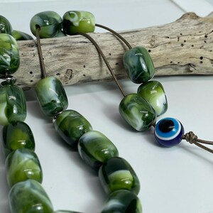 Op de afbeelding: Een groene en witte gebedskralenketting met een blauwe en witte boze oog-hanger. De kralen zijn gemaakt van gemarmerd materiaal en zijn geregen aan een bruin koord.