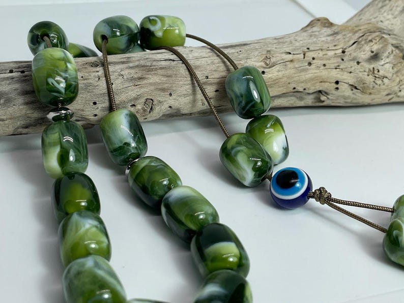 Op de afbeelding: Een groene en witte gebedskralenketting met een blauwe en witte boze oog-hanger. De kralen zijn gemaakt van gemarmerd materiaal en zijn geregen aan een bruin koord.