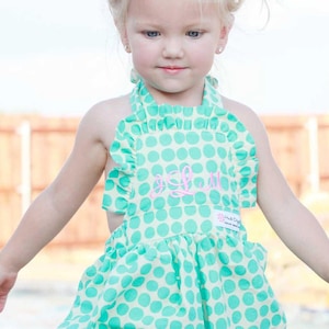 May include: A little girl wearing a green and white polka dot sunsuit with a pink embroidered letter "L". The sunsuit has a ruffled halter top and a gathered skirt. The sunsuit is made by Haute Diggity.