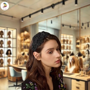 May include: A black floral headband featuring three large fabric flowers and small beaded accents. The headband is worn by a person with long brown hair. The background shows a hair salon with wigs on display.
