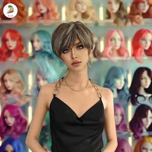 May include: A woman with short, gray and brown hair is wearing a black halter top with a gold chain detail. She is standing in front of a wall with many colorful wigs.