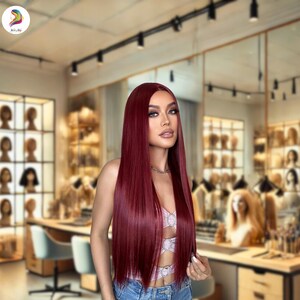 May include: A woman with long, straight, burgundy-colored hair. She is wearing a pink top and blue jeans. The background is a hair salon with wigs on display.