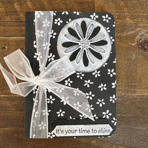 May include: A black notebook with a white floral pattern and a silver daisy design. The notebook has a white ribbon tied around it and the text "It's your time to shine" on the front.