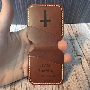 May include: A brown leather wallet featuring a cross symbol and the inscription "I AM The Way The Truth The Life". The wallet has a stitched border and card slots.