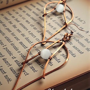 May include: A pair of handcrafted copper wire earrings with a teardrop design, each featuring a white, round bead. The earrings are displayed on an open book with faded text, showcasing a vintage aesthetic.