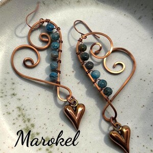 May include: A pair of copper wire earrings shaped like open hearts with blue and grey stone beads wrapped along one side. Each earring features a small, solid copper heart charm dangling from the bottom. The text Marokel is visible.