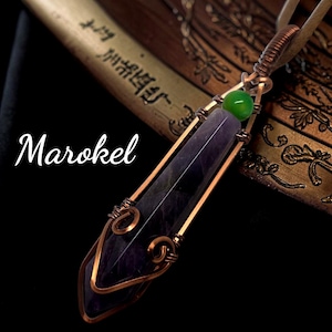 May include: A pendant featuring a long, faceted purple amethyst crystal point encased in copper wire wrapping with decorative swirls. A small, round, bright green bead sits above the crystal. The word Marokel is written in white script.