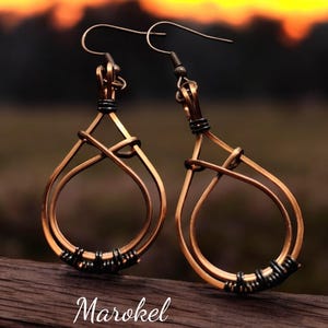 May include: A pair of copper wire earrings with a teardrop shape and black wire accents. The earrings are hanging from a wooden surface.