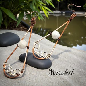 May include: A pair of handcrafted copper and silver-toned earrings. Each earring features a teardrop shape with a white stone bead and a series of silver-toned circles at the base. The earrings are displayed on a gray surface with the word "Marokel".