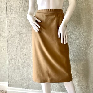 May include: A tan-coloured, knee-length skirt displayed on a mannequin. The skirt has a straight cut and a simple design, with a waistband and a subtle seam down the back. The fabric appears to be a smooth, woven material.