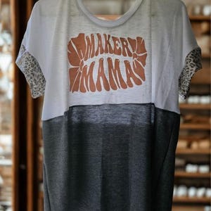 Puede incluir: Camiseta de dos tonos con una sección superior blanca y una sección inferior gris oscuro. Las mangas tienen un estampado de leopardo. La camiseta presenta el texto "MAKER MAMAS" en una fuente estilizada de color óxido.