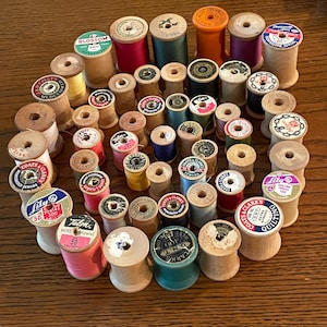 May include: A collection of vintage wooden spools of thread in various colors, including red, blue, yellow, and green. Some spools have labels with text like "COATS" and "Lily". The spools are arranged on a wooden surface.