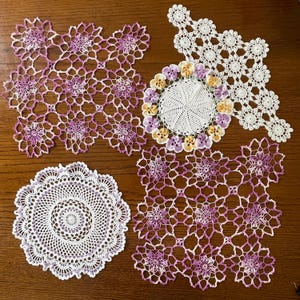 Può includere: Una collezione di centrini all'uncinetto fatti a mano in varie forme e dimensioni. I centrini presentano intricati motivi traforati e sono principalmente bianchi con accenti di viola e giallo. La disposizione comprende disegni rotondi e quadrati, che mostrano la delicata maestria artigianale.