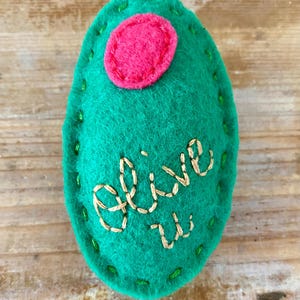 May include: A handmade felt ornament in the shape of an olive. The green felt olive has a pink circle on top and is embroidered with the words "olive u" in gold thread. The edges are stitched with green thread.
