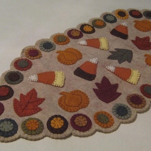May include: A beige felt table runner with a scalloped edge, decorated with autumn-themed appliqués. The design includes pumpkins, leaves, and candy corn in shades of orange, brown, and yellow. The runner is approximately 36 inches long.
