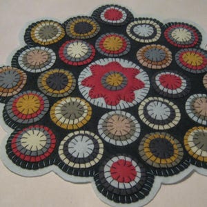 May include: A handmade, circular rug with a scalloped edge. The rug features a dark background with numerous colorful circles in shades of red, yellow, gray, and beige. A large red flower is in the center. The rug is likely made of felt.