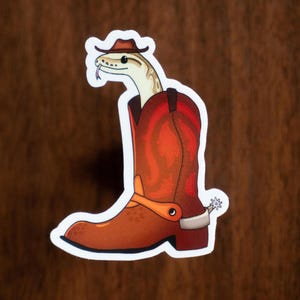 May include: A sticker featuring a cartoon snake wearing a cowboy hat and emerging from a brown cowboy boot. The boot has orange and brown tones, with a spur on the heel. The snake has a cream-colored body and a forked tongue.