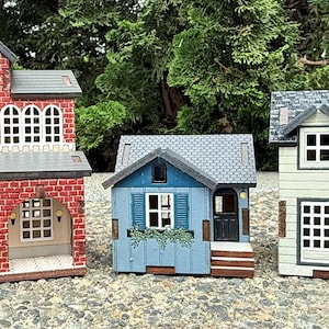 Puede incluir: Tres casas modelo en miniatura. La más grande es un edificio de ladrillo rojo con una torre de reloj. En el medio hay una casa azul con molduras blancas y un tejado gris. La tercera casa es blanca con un tejado gris.