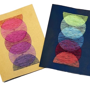 May include: Two notebooks with abstract designs. One is tan with a stacked design of pink, purple, and blue shapes. The other is navy blue with a similar design in red, purple, blue, and green.