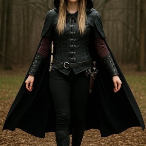 May include: A woman in a black hooded cloak and leather corset walks through a forest. She is wearing black pants and boots, and has a dagger strapped to her leg.