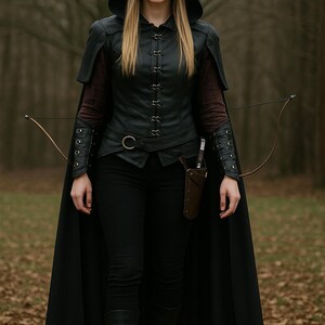May include: A woman in a black hooded cloak and leather vest with a bow and arrow. She is standing in a forest setting.