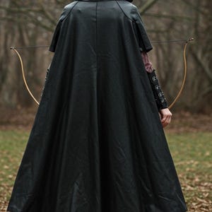 May include: A black leather hooded cape with a long, flowing design. The cape has a high collar and a pointed hood. The person wearing the cape is holding a bow and arrow.