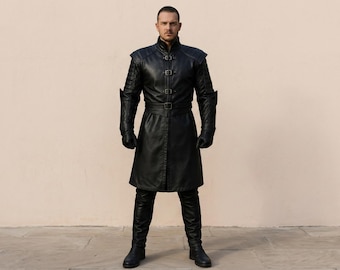 May include: A black leather-style long coat featuring a high collar, three front buckle closures, and a matching waist belt. The garment includes quilted shoulder and sleeve detailing, long sleeves, and a knee-length hem, paired with black trousers.