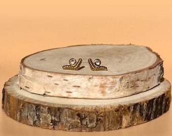 May include: A pair of gold-colored stud earrings, each featuring a small, clear gemstone. The earrings are displayed on a rustic, round wooden surface with a light brown background. The earrings have a unique, curved design.
