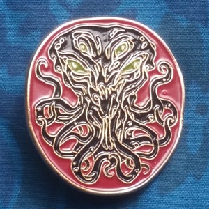 May include: A red and gold-colored pin with a black and green illustration of a tentacled creature with multiple eyes. The pin is about the size of a US quarter.
