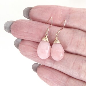May include: A pair of teardrop-shaped earrings with faceted, pale pink gemstones. The gemstones are suspended from gold-colored wire hooks, with gold wire wrapped around the top of each stone. The earrings are displayed against a light background.