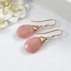 May include: A pair of drop earrings featuring faceted, teardrop-shaped pink gemstones. The gemstones are wire-wrapped in gold-colored metal and suspended from gold-colored ear wires. The earrings are displayed on a white surface.