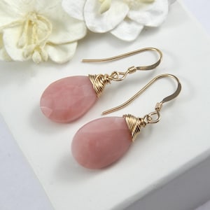 May include: A pair of gold-colored drop earrings with teardrop-shaped, faceted pink gemstones. The gemstones are wrapped with gold wire and attached to gold-colored ear wires. The earrings are displayed on a white surface.