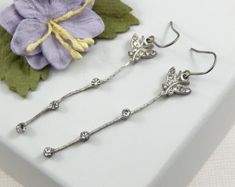 May include: Silver-toned dangle earrings featuring a butterfly design at the top, embellished with clear crystals. The earrings have a textured, linear design with three additional crystals dangling below the butterfly. The earrings are designed with a hook closure.