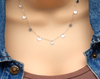 May include: A silver chain necklace with small, round silver charms hanging from it. The necklace is on a model wearing a brown shirt and a denim jacket.