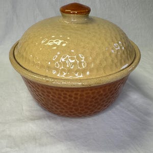 May include: A brown and beige ceramic casserole dish with a lid. The dish has a textured, hammered finish. The lid has a small knob on top.