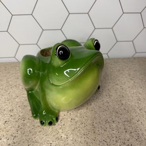 May include: A green ceramic frog planter with black eyes and a wide, toothy grin. The frog is sitting on a light brown surface.