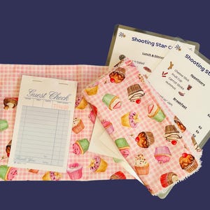 May include: A pink and white checkered server's apron with a cupcake pattern, a teal pencil, and a guest check. Two menu cards for "Shooting Star Cafe" are visible. The apron has a pink strap.