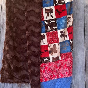 May include: A brown faux fur blanket with a patchwork quilt on top. The quilt is made of red, blue, and white fabric with a paisley pattern. The quilt has a variety of images including horses, saddles, and cowboy boots. The text "Saddle Up" is visible on the quilt.