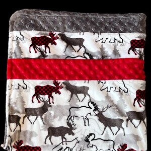 May include: A soft, plush baby blanket with a white background featuring a gray and red plaid pattern. The blanket has a red and gray polka dot border and a red polka dot stripe in the center. The blanket features a woodland animal print with moose, deer, and bears.
