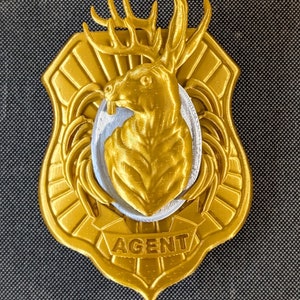 Könnte beinhalten: Ein goldfarbenes Abzeichen mit einem Hirschkopf in der Mitte, vor einem silbernen Oval. Das Abzeichen ist schildförmig und trägt am unteren Rand das Wort "AGENT". Der Hintergrund ist ein dunkelgraues, strukturiertes Gewebe.