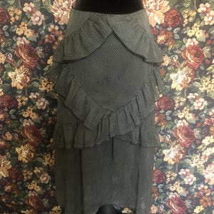 Op de afbeelding: Een zwart-witte polkadot rok met gelaagde ruches. De rok wordt getoond op een zwarte mannequin. De rok heeft een hoge taille en een uitlopend silhouet. De achtergrond is een bloemenpatroon.