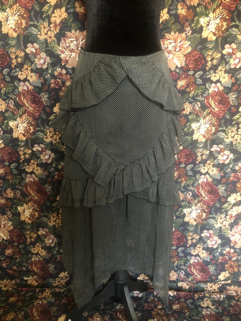 Op de afbeelding: Een zwart-witte polkadot rok met gelaagde ruches. De rok wordt getoond op een zwarte mannequin. De rok heeft een hoge taille en een uitlopend silhouet. De achtergrond is een bloemenpatroon.
