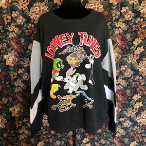 May include: A black long-sleeve sweatshirt featuring the red text "LOONEY TUNES" and a graphic print of various characters including Bugs Bunny, Tweety Bird, Marvin the Martian, and Wile E. Coyote. The sleeves have grey and white color-blocked panels.