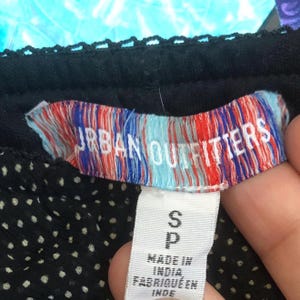 Op de afbeelding: Close-up van een zwart kledingstuk met een wit stippenpatroon. Het label leest "URBAN OUTFITTERS" in witte letters tegen een rood, wit en blauw gestreepte achtergrond. Onder het label staat een wit label met de maat "S" en "P", en de tekst "MADE IN INDIA".