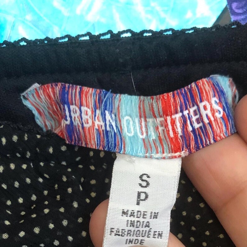 Op de afbeelding: Close-up van een zwart kledingstuk met een wit stippenpatroon. Het label leest "URBAN OUTFITTERS" in witte letters tegen een rood, wit en blauw gestreepte achtergrond. Onder het label staat een wit label met de maat "S" en "P", en de tekst "MADE IN INDIA".