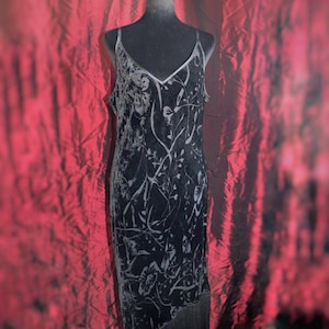 May include: Black velvet slip dress with a floral pattern and fringe hem. The dress features a V-neckline with a silver trim and spaghetti straps. The dress is displayed on a mannequin against a red backdrop.